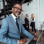Professional man in a blue suit working on a laptop in a modern office setting.